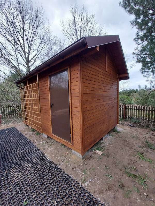 Mały, drewniany budynek gospodarczy z brązowymi drzwiami i kratownicą z boku, ustawiony na podłożu z geokraty.