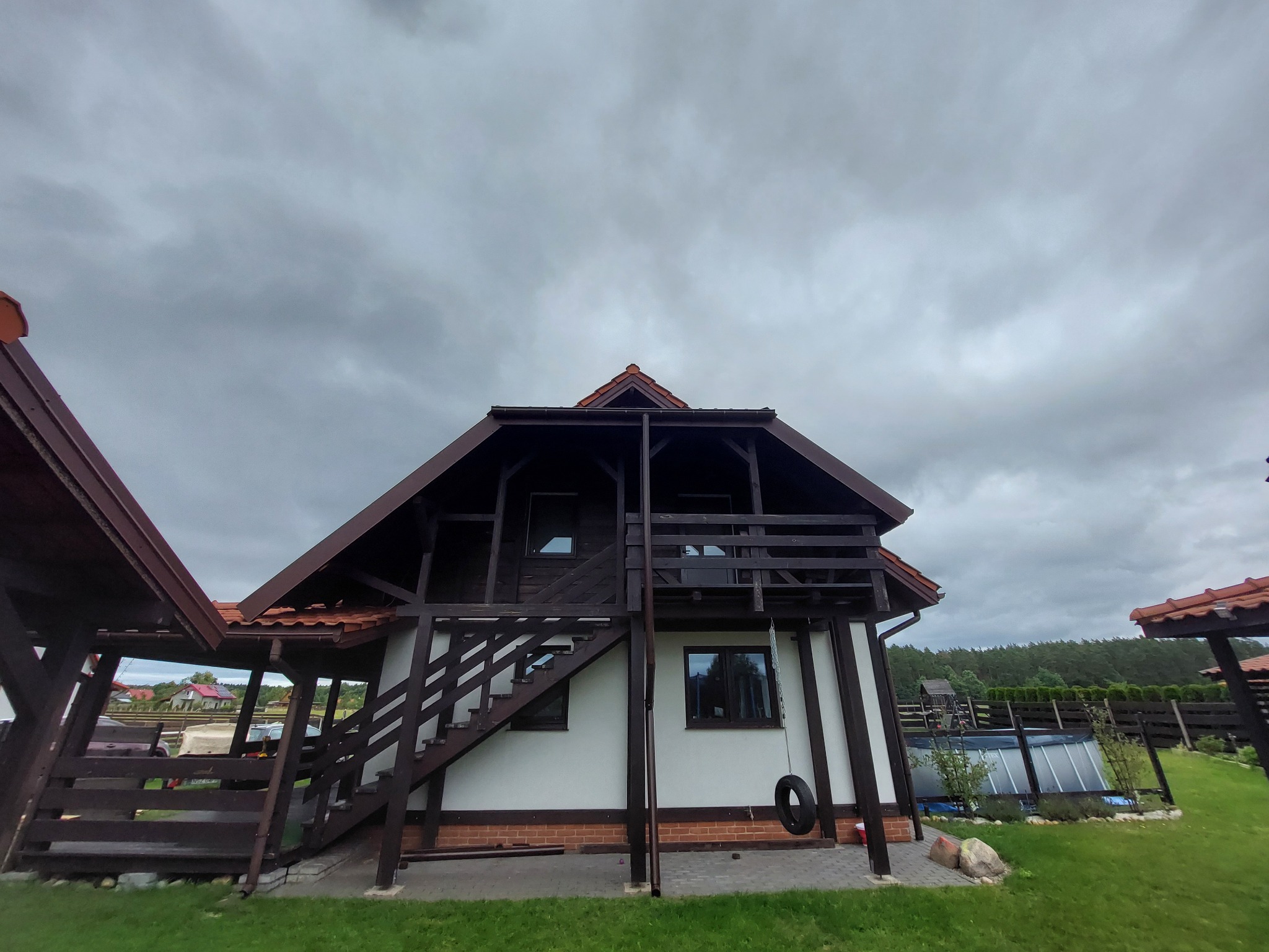 Dwukondygnacyjny dom z białymi ścianami i ciemnobrązową drewnianą konstrukcją, zewnętrznymi schodami i balkonem, z oponą zawieszoną na łańcuchu, otoczony zielonym trawnikiem i ogrodzeniem, na tle...