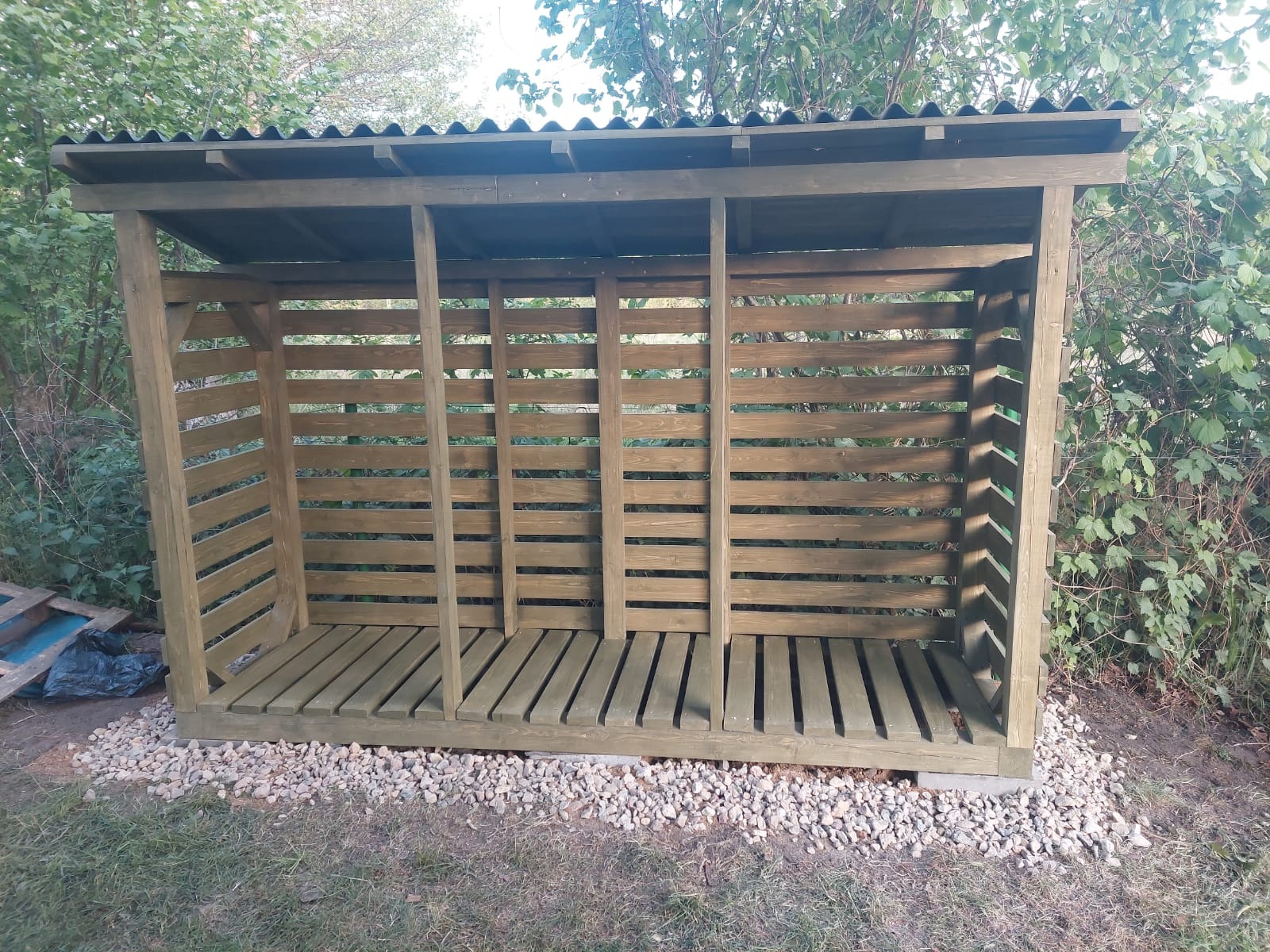 Drewniana konstrukcja z zadaszeniem, prawdopodobnie drewutnia, z poziomymi szczebelkami, ustawiona na podłożu wysypanym drobnymi kamieniami, w tle zieleń drzew i krzewów.