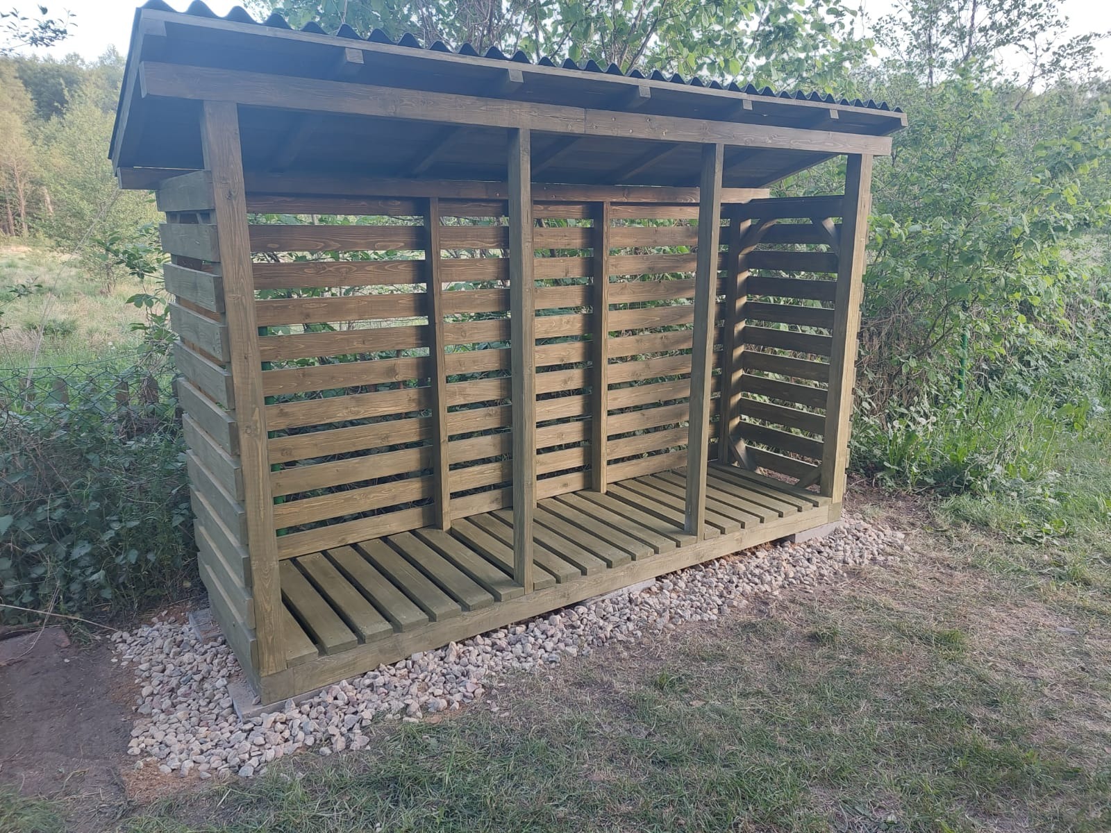 Drewniana wiata na drewno opałowe z ażurowymi ścianami, ustawiona na fundamencie z kamieni, z zielonym tłem drzew i krzewów.