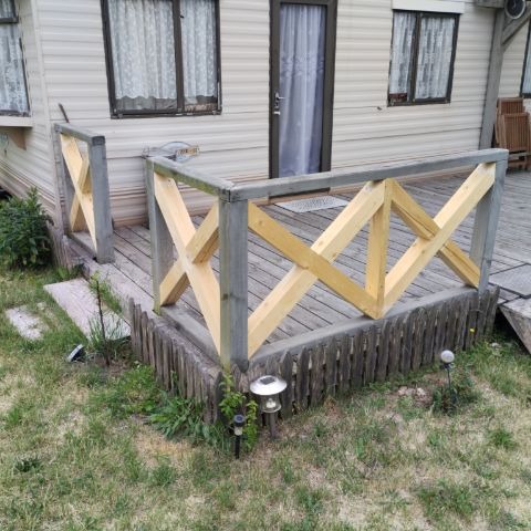 Drewniany taras z balustradą o wzorze krzyżowym, przylegający do domu mobilnego, otoczony trawnikiem z lampkami solarnymi.