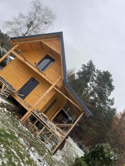 Szkielet domu w trakcie budowy, drewniana konstrukcja z widocznymi belkami, szary dach, częściowo ośnieżone otoczenie, drzewa w tle.