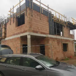 Dwupiętrowy budynek w trakcie budowy z czerwonej cegły, widoczne drewniane rusztowania i betonowe elementy konstrukcyjne, na pierwszym planie zaparkowany szary samoch&oacute;d z otwartym bagażnikiem.