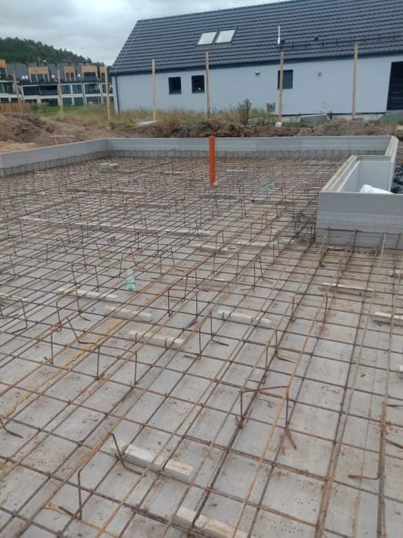 Przygotowanie zbrojenia płyty fundamentowej pod budynek, widoczne siatki z prętów stalowych i szalunek obwodowy, w tle nowo powstające domy.