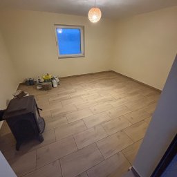 Oskar Romanowski - Pusta przestrzeń z nową podłogą z paneli imitujących drewno, grzejnikiem typu koza i materiałami budowlanymi przy ścianie pod oknem, oświetlona wiszącą lampą z plecionki.