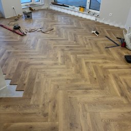 Oskar Romanowski - Układanie parkietu w jodełkę w jasnym pomieszczeniu z dużymi oknami, widoczne narzędzia pracy i niedokończony fragment podłogi przy ścianie.