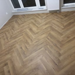 Oskar Romanowski - Ułożona pod kątem 45 stopni podłoga z paneli w jodełkę francuską, widok z góry na jasne, pomalowane ściany i biały grzejnik pod oknem.