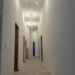 Oskar Romanowski - Długi korytarz w trakcie remontu, świeżo pomalowane na biało ściany, heksagonalna podłoga i surowe ościeżnice drzwiowe.