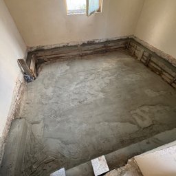 Oskar Romanowski - Przygotowanie posadzki w remontowanym pomieszczeniu z oknem dachowym; widoczne drewniane elementy szalunku i świeża wylewka betonowa.