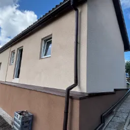 Odnowiona elewacja budynku z dwukolorowym tynkiem, nowymi oknami i systemem rynnowym. Na pierwszym planie skrzynki, błękitne niebo w tle.