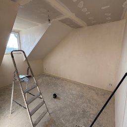 Oskar Romanowski - Poddasze po szpachlowaniu, gotowe do malowania. Widoczna drabina, wiadro z wałkiem i laserowy niwelator. Skosy ścian i sufitu, okno dachowe.
