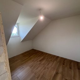 Oskar Romanowski - Pokój na poddaszu po remoncie: drewnopodobne panele ułożone na podłodze, świeżo pomalowane ściany na biało, okno w skosie, surowa ściana z lewej strony.