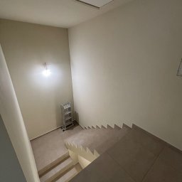 Oskar Romanowski - Wykończone schody wewnętrzne z brązowymi płytkami, widok z góry. Na ścianie widoczna drabina aluminiowa i niedokończone oświetlenie. Sufit z klapą włazową.