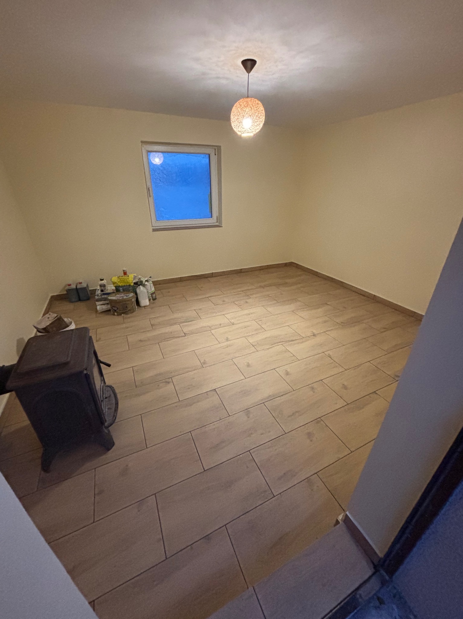 Pusta przestrzeń z nową podłogą z paneli imitujących drewno, grzejnikiem typu koza i materiałami budowlanymi przy ścianie pod oknem, oświetlona wiszącą lampą z plecionki.