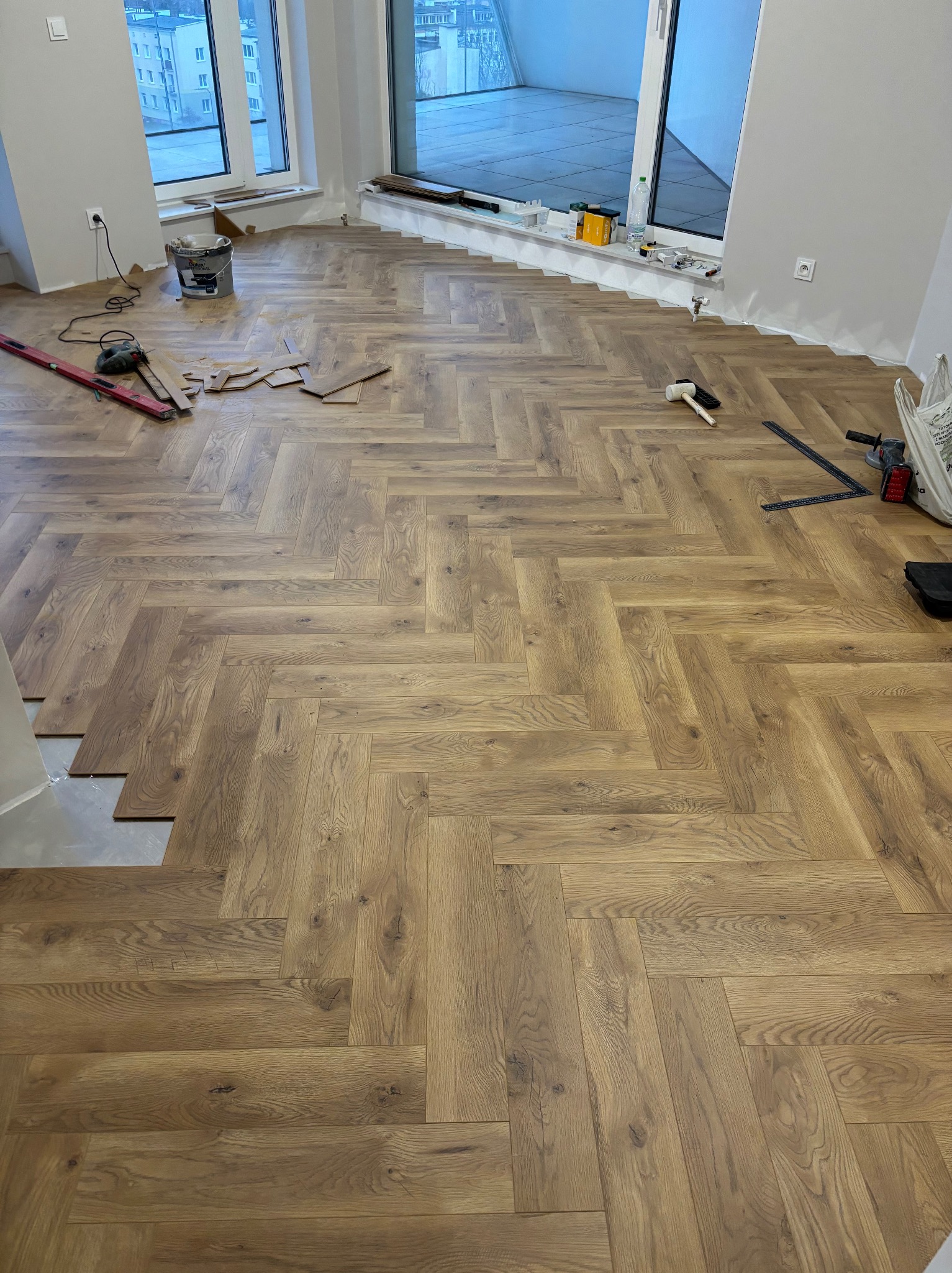 Układanie parkietu w jodełkę w jasnym pomieszczeniu z dużymi oknami, widoczne narzędzia pracy i niedokończony fragment podłogi przy ścianie.