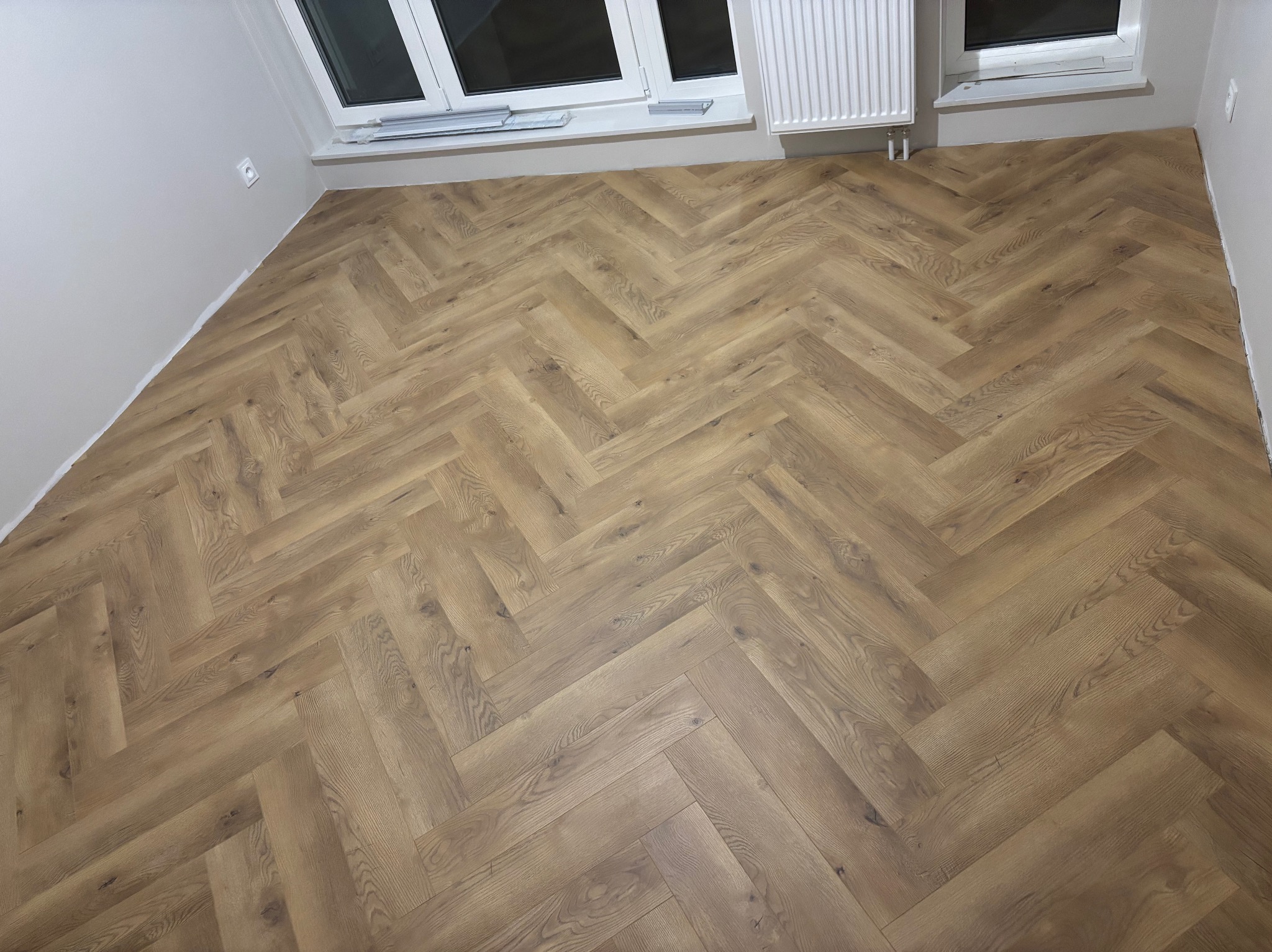 Ułożona pod kątem 45 stopni podłoga z paneli w jodełkę francuską, widok z góry na jasne, pomalowane ściany i biały grzejnik pod oknem.