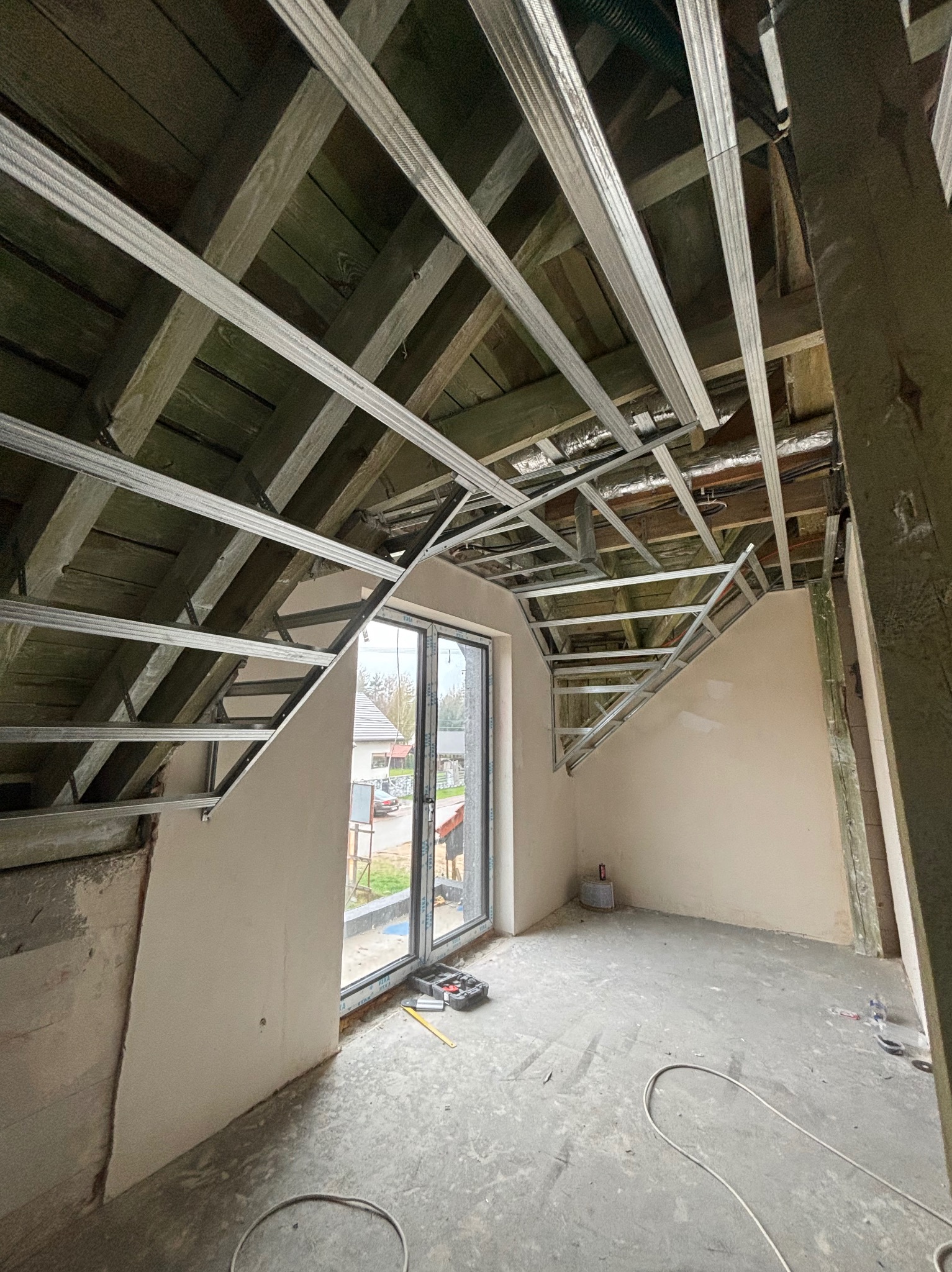 Adaptacja poddasza: metalowa konstrukcja sufitu podwieszanego na drewnianej więźbie, ściany częściowo wyszpachlowane, okno dachowe, narzędzia na podłodze.