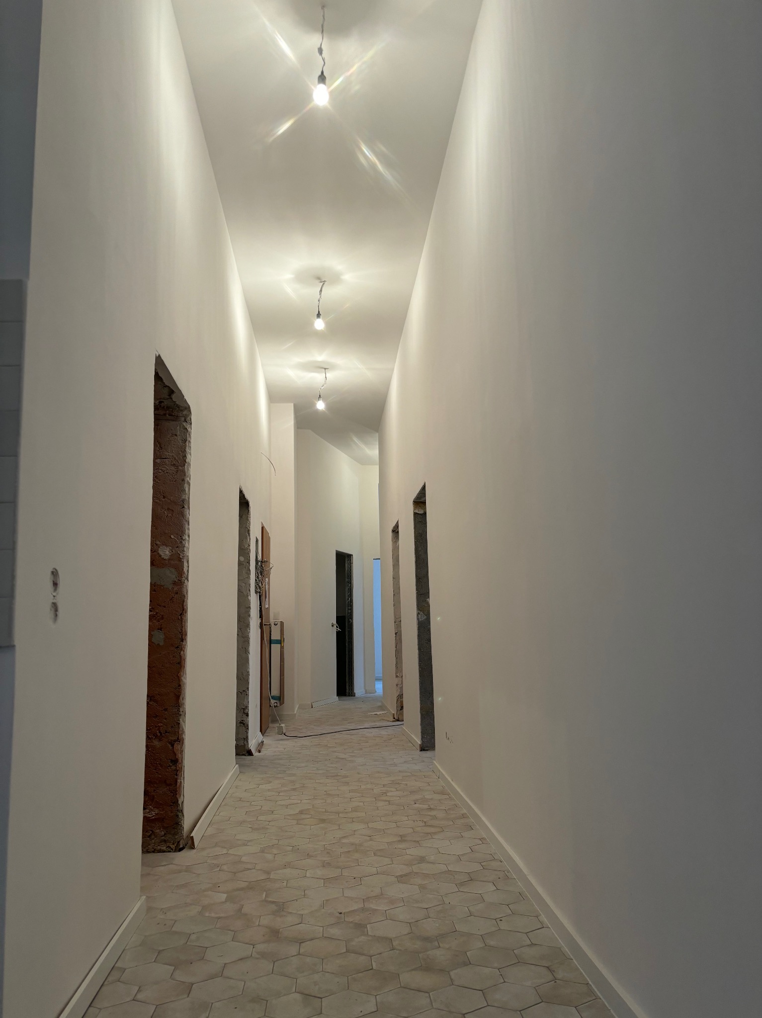 Długi korytarz w trakcie remontu, świeżo pomalowane na biało ściany, heksagonalna podłoga i surowe ościeżnice drzwiowe.