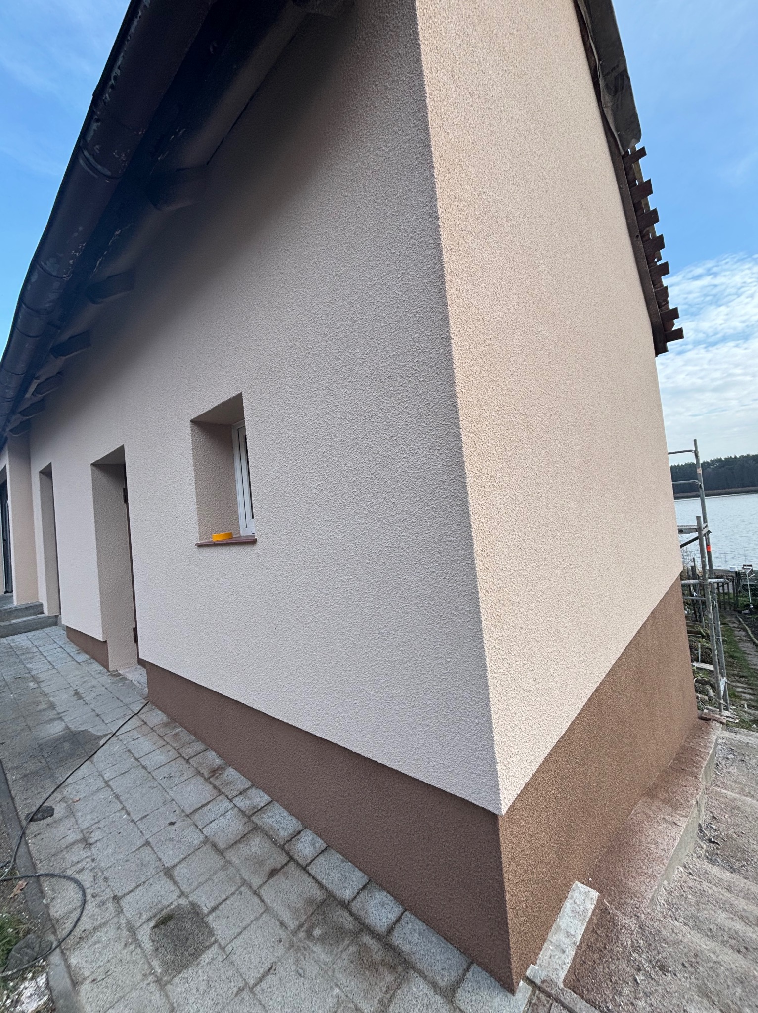 Elewacja budynku z oknem, wykończona jasnym tynkiem strukturalnym i brązowym cokołem, widok z zewnątrz na tle błękitnego nieba i jeziora, z widocznymi rusztowaniami przy ścianie szczytowej.