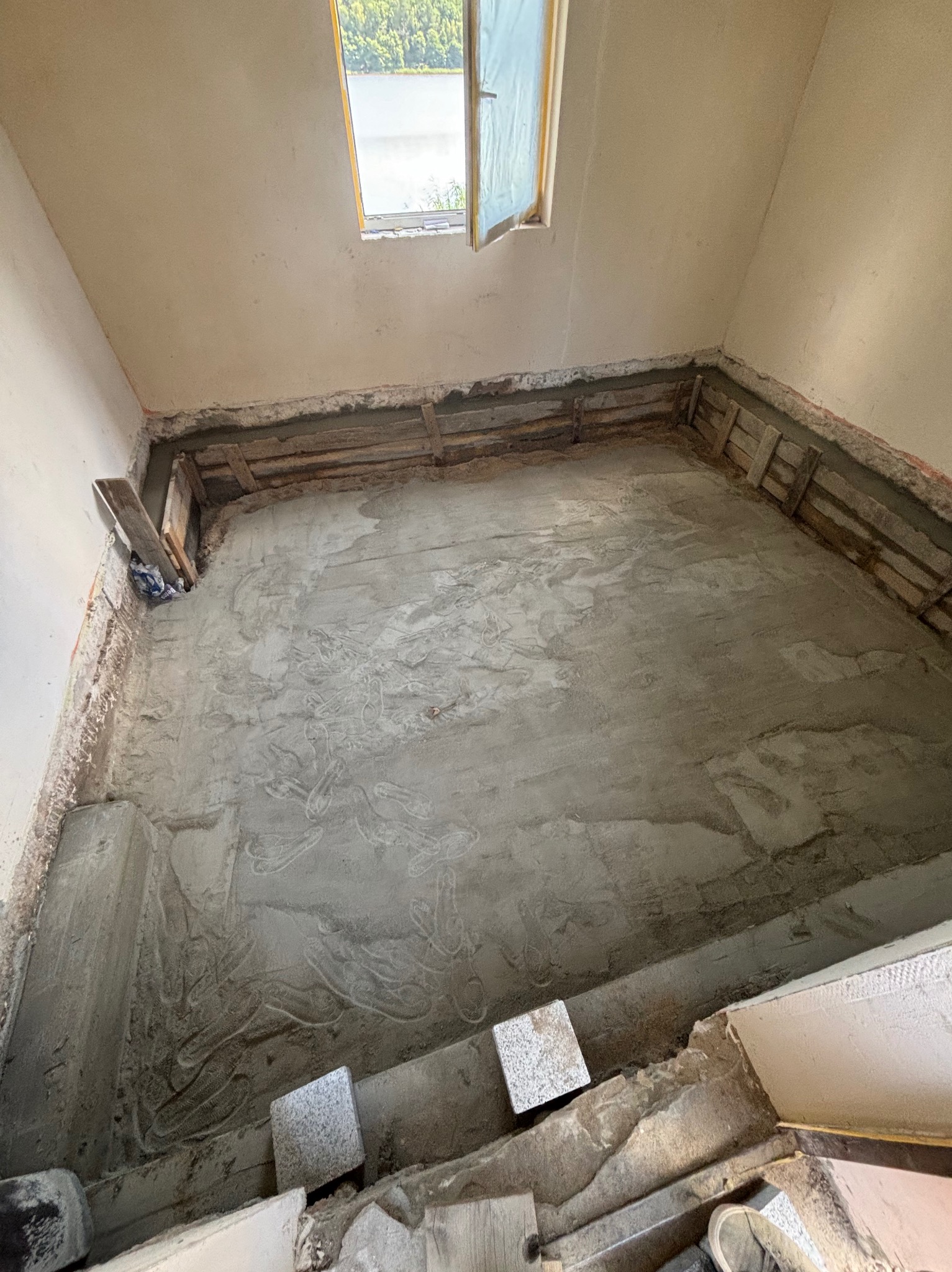 Przygotowanie posadzki w remontowanym pomieszczeniu z oknem dachowym; widoczne drewniane elementy szalunku i świeża wylewka betonowa.