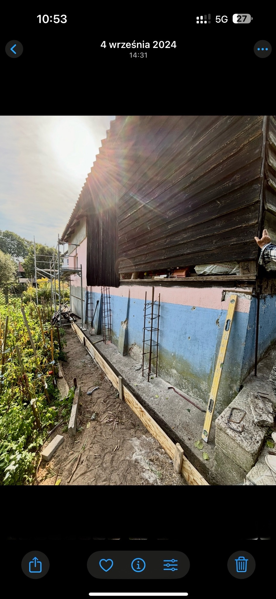 Stodoła w trakcie rozbudowy: widoczne fundamenty z deskowaniem, zbrojenia, poziomica oparta o ścianę budynku i rusztowania w tle. Prace ziemne w toku.