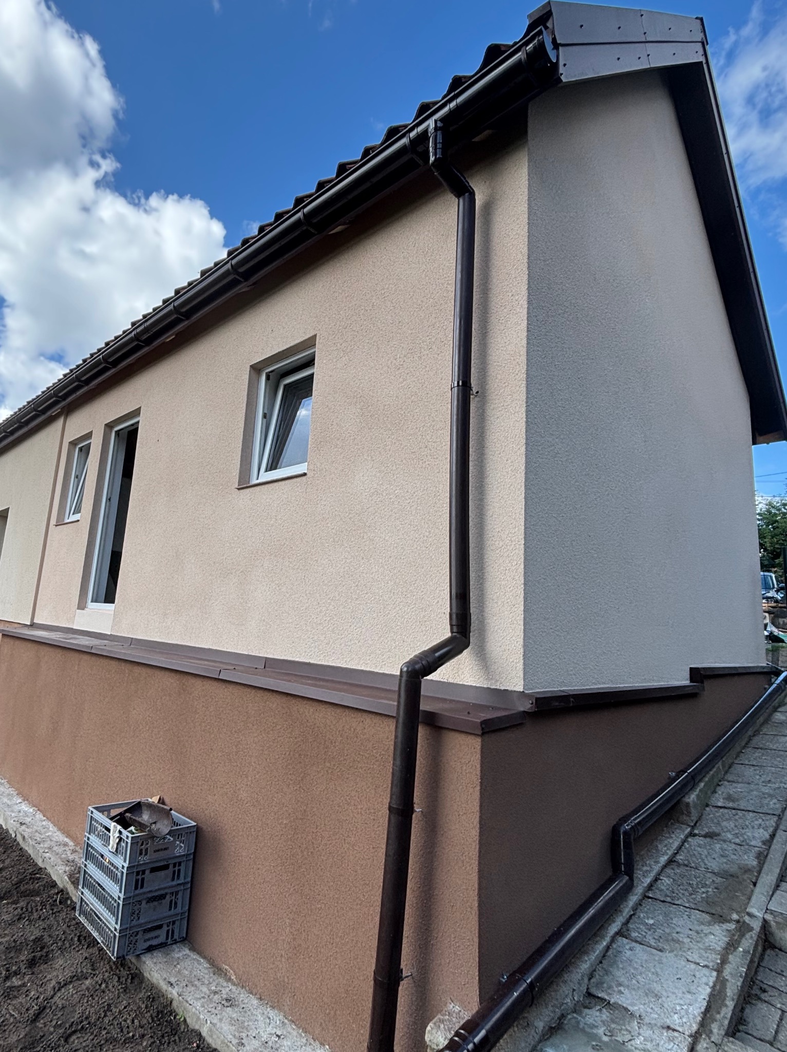 Odnowiona elewacja budynku z dwukolorowym tynkiem, nowymi oknami i systemem rynnowym. Na pierwszym planie skrzynki, błękitne niebo w tle.