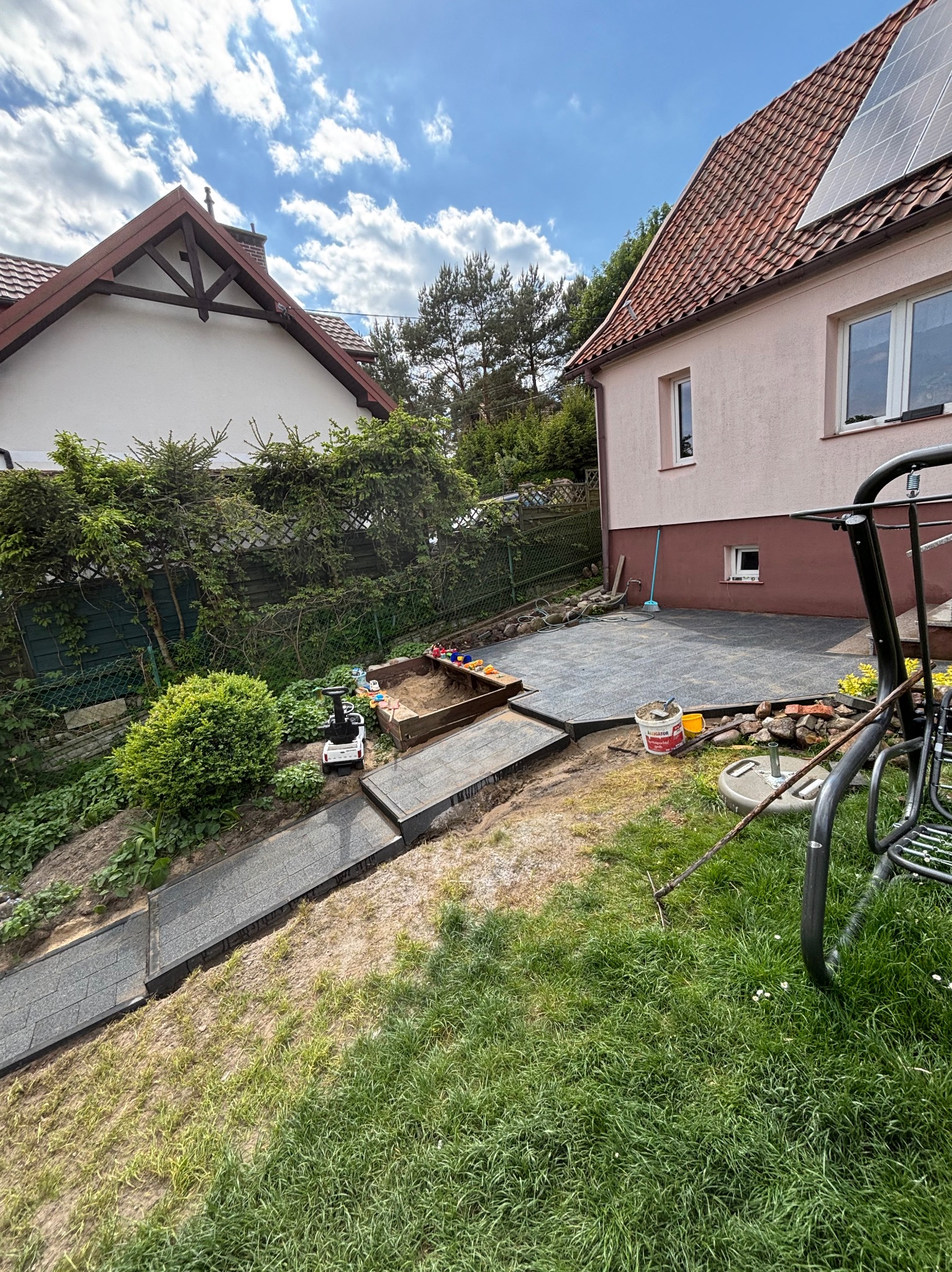 Taras z szarych płyt na nierównym terenie, z widoczną ścieżką z płyt i piaskownicą. W tle dom z panelami słonecznymi i bujna zieleń.