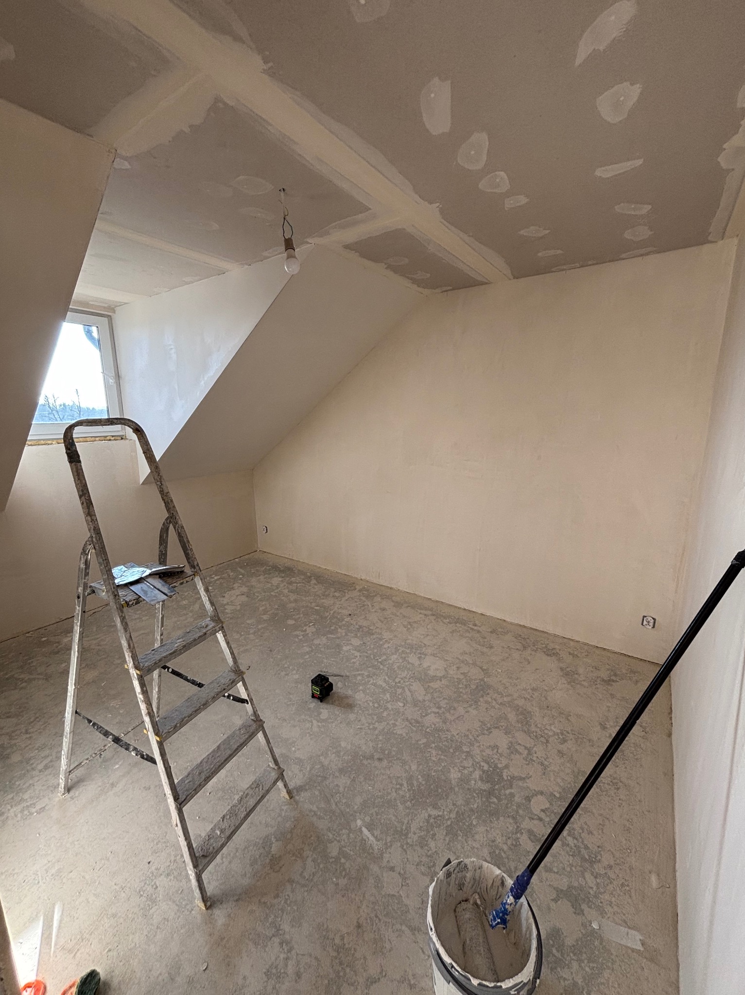 Poddasze po szpachlowaniu, gotowe do malowania. Widoczna drabina, wiadro z wałkiem i laserowy niwelator. Skosy ścian i sufitu, okno dachowe.