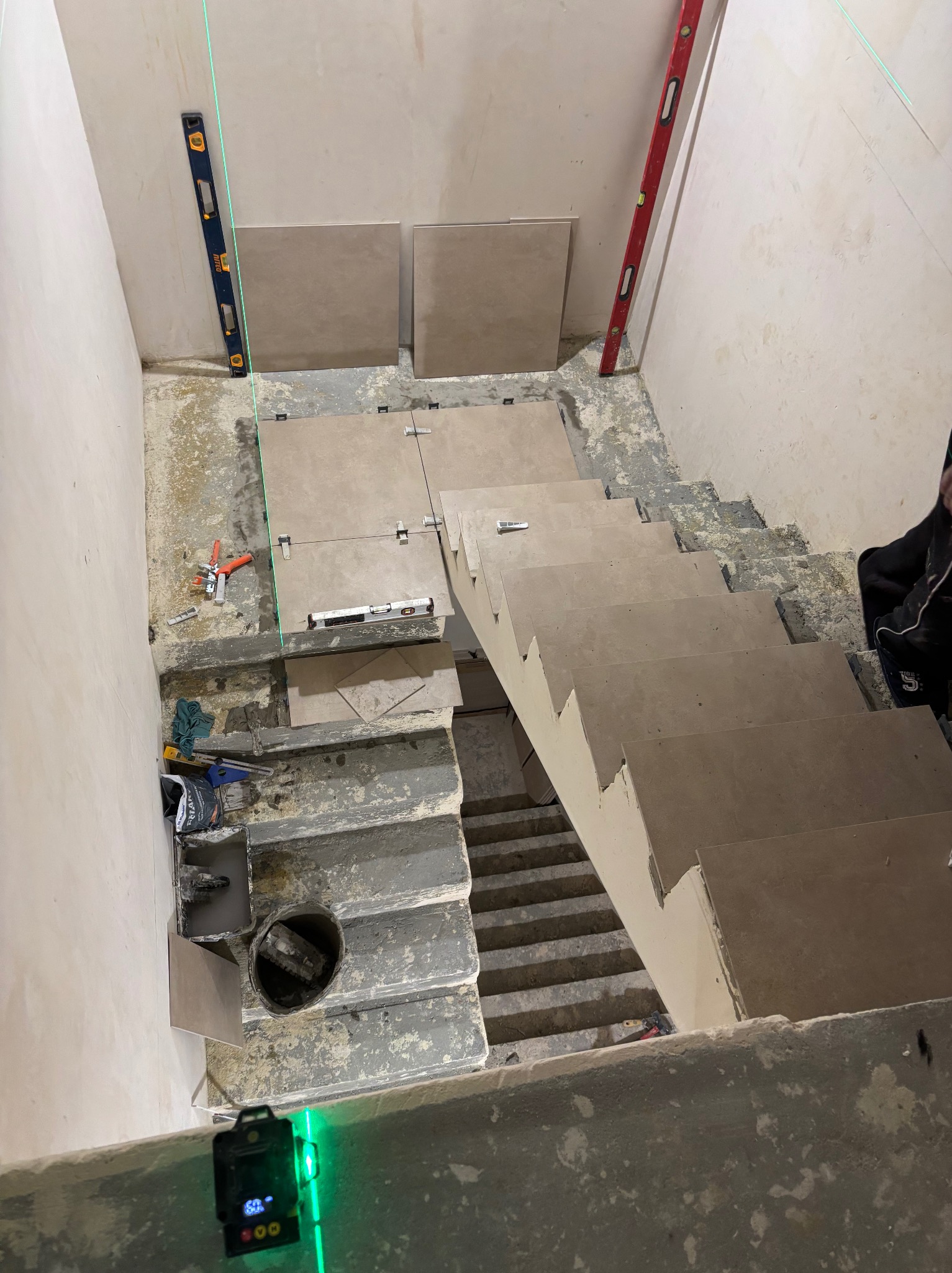 Montaż płytek na schodach z użyciem poziomicy laserowej. Widoczne płytki, poziomice, narzędzia glazurnicze i fragment schodów w trakcie wykańczania.