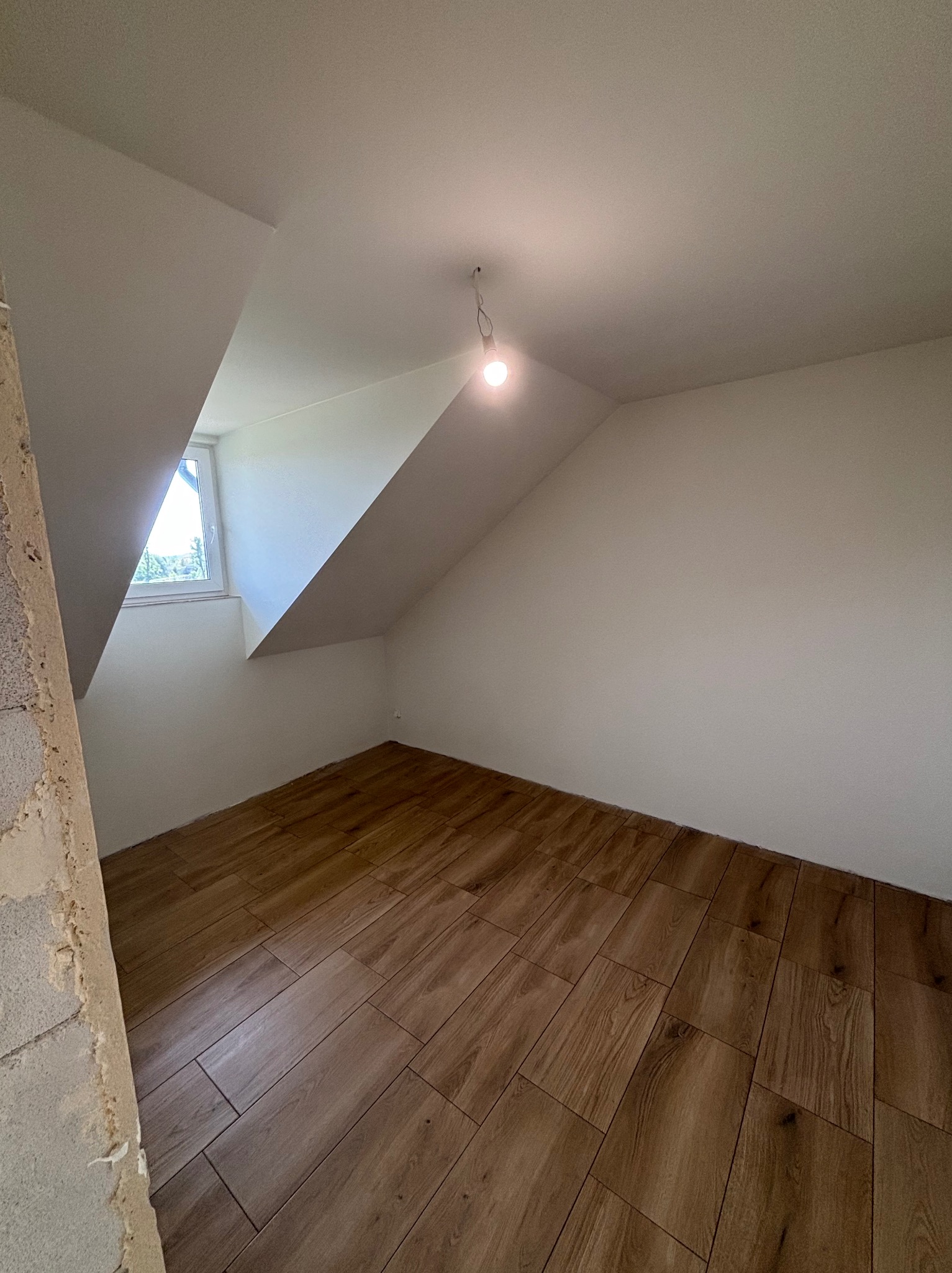 Pokój na poddaszu po remoncie: drewnopodobne panele ułożone na podłodze, świeżo pomalowane ściany na biało, okno w skosie, surowa ściana z lewej strony.