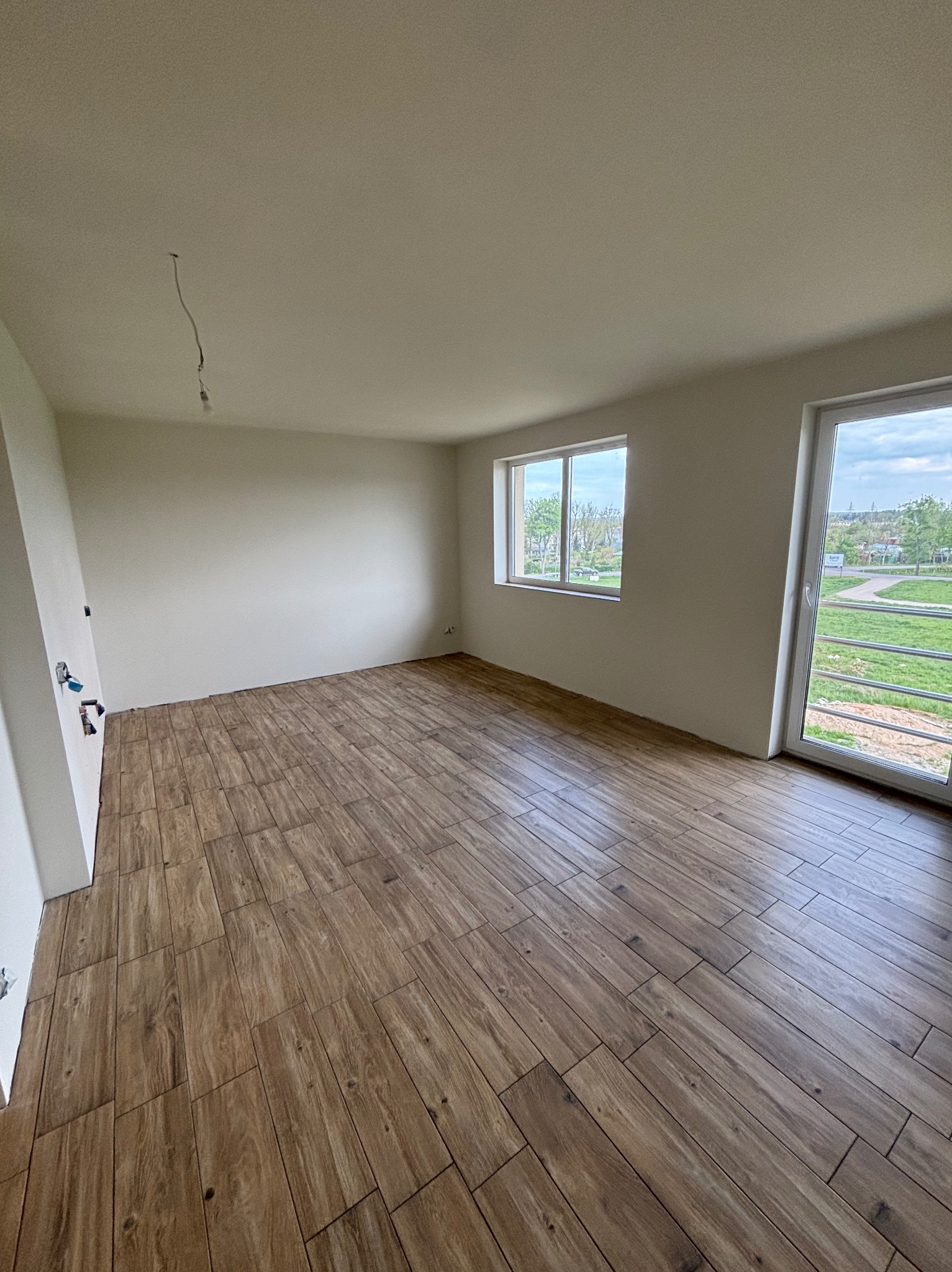 Jasne, puste wnętrze z nową, drewnopodobną podłogą. Widoczne okno i drzwi balkonowe. Ściany przygotowane do malowania, z widocznymi przewodami elektrycznymi.