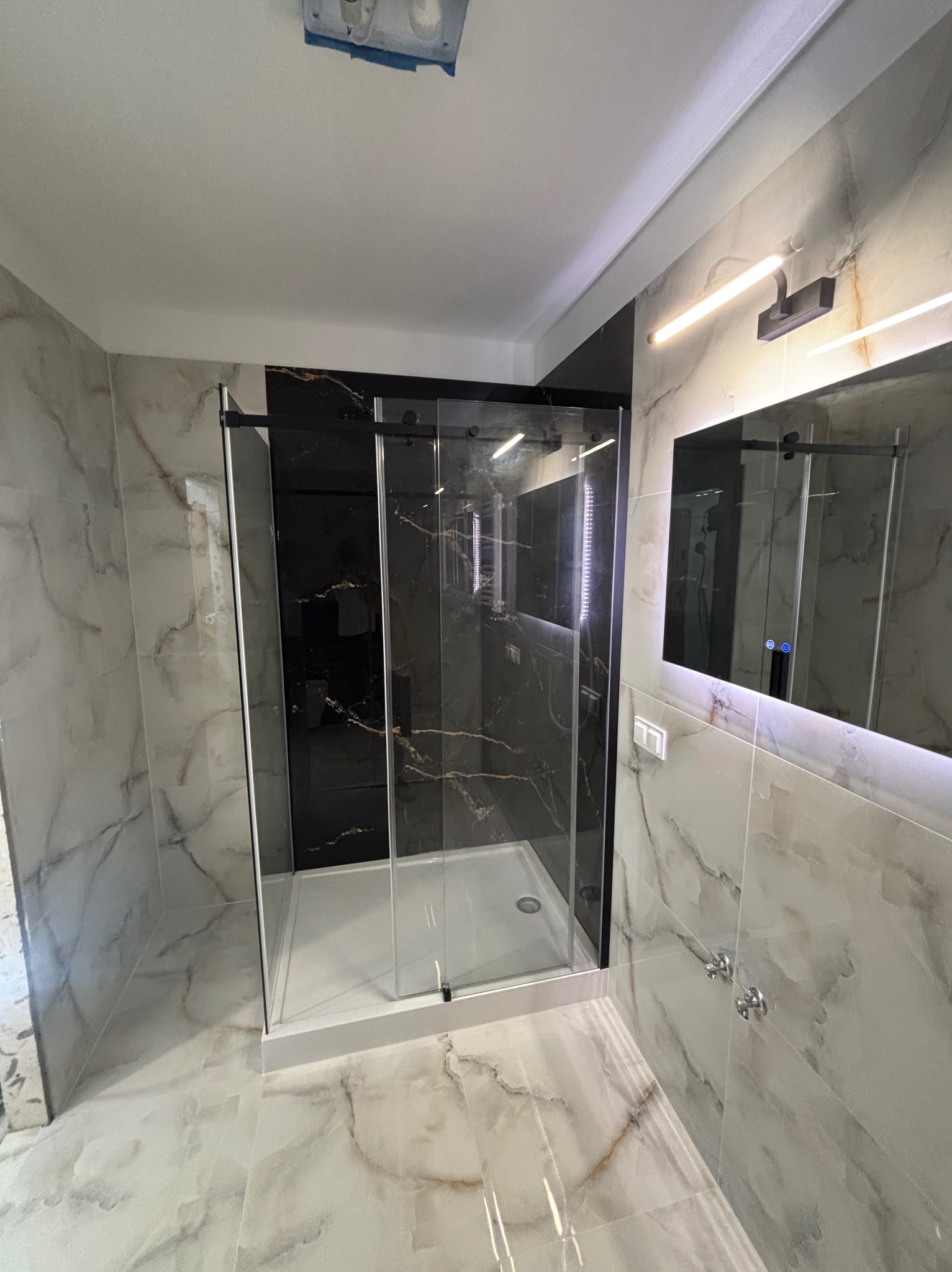 Nowoczesna kabina prysznicowa z czarnymi elementami i marmurowymi płytkami w wyremontowanej łazience. Lustro z podświetleniem LED. Widok z perspektywy.