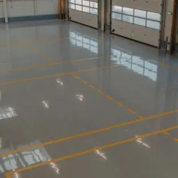 Błyszcząca, szara podłoga w hali przemysłowej z wyznaczonymi żółtymi liniami, widok z góry na otwarte bramy segmentowe.