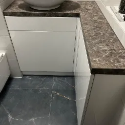 Nowoczesna łazienka z białą umywalką nablatową, brązowym blatem imitującym marmur, białymi szafkami pod umywalką i ciemną, marmurkową podłogą.