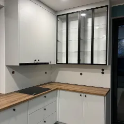 Nowoczesna kuchnia z szarymi frontami, drewnianym blatem i przeszklonymi szafkami wiszącymi z czarnymi ramami, biała ściana z płytkami ułożonymi w jodełkę, minimalistyczne czarne uchwyty.