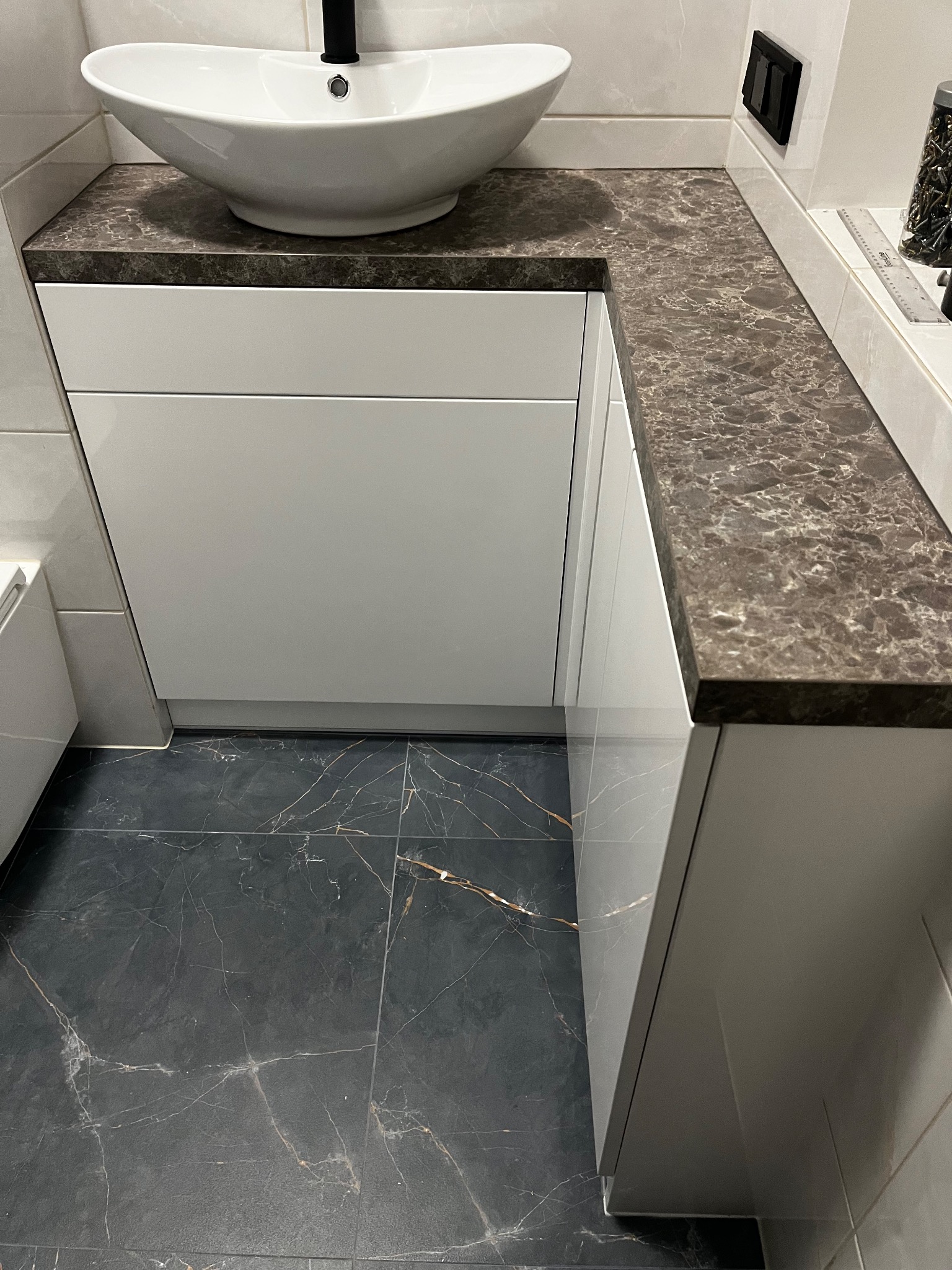 Nowoczesna łazienka z białą umywalką nablatową, brązowym blatem imitującym marmur, białymi szafkami pod umywalką i ciemną, marmurkową podłogą.