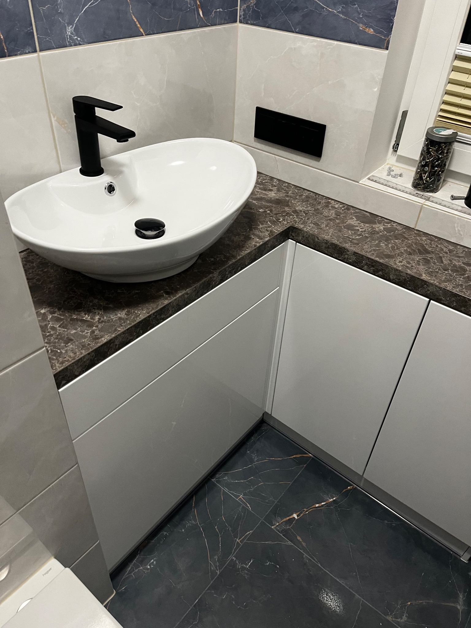 Nowoczesna łazienka z białą owalną umywalką, czarną baterią, blatem imitującym marmur i białymi szafkami pod umywalką, uzupełniona marmurową podłogą i płytkami ściennymi.