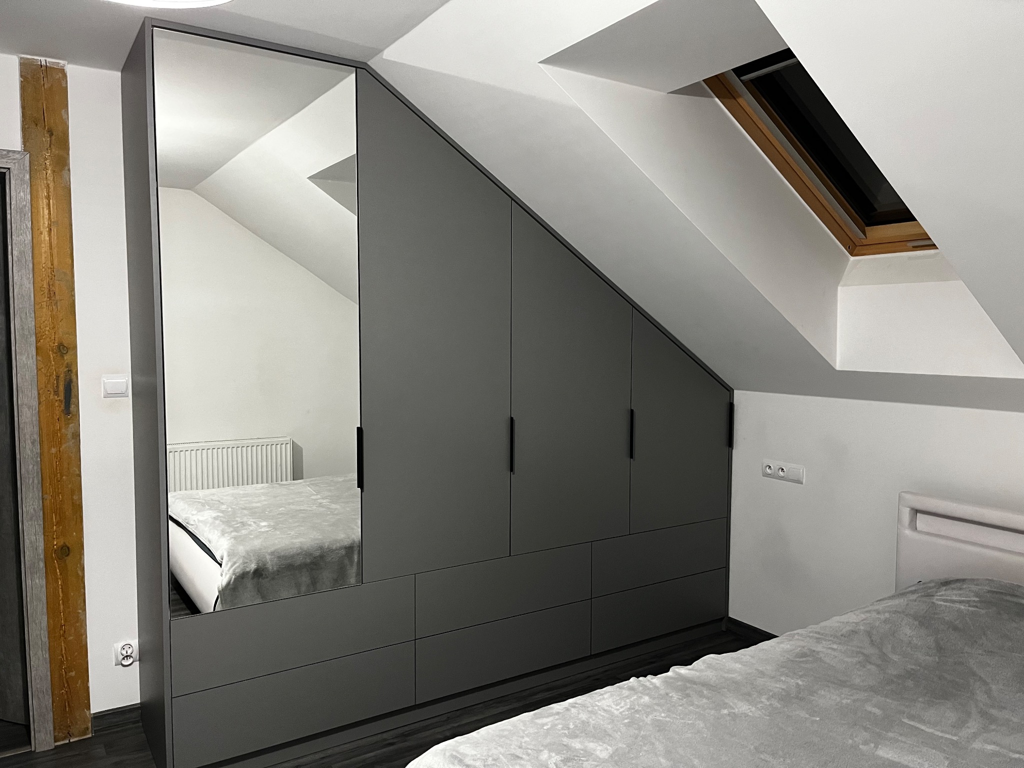 Szara szafa wnękowa z lustrem, dopasowana do skosu poddasza, widoczna w sypialni z oknem dachowym.