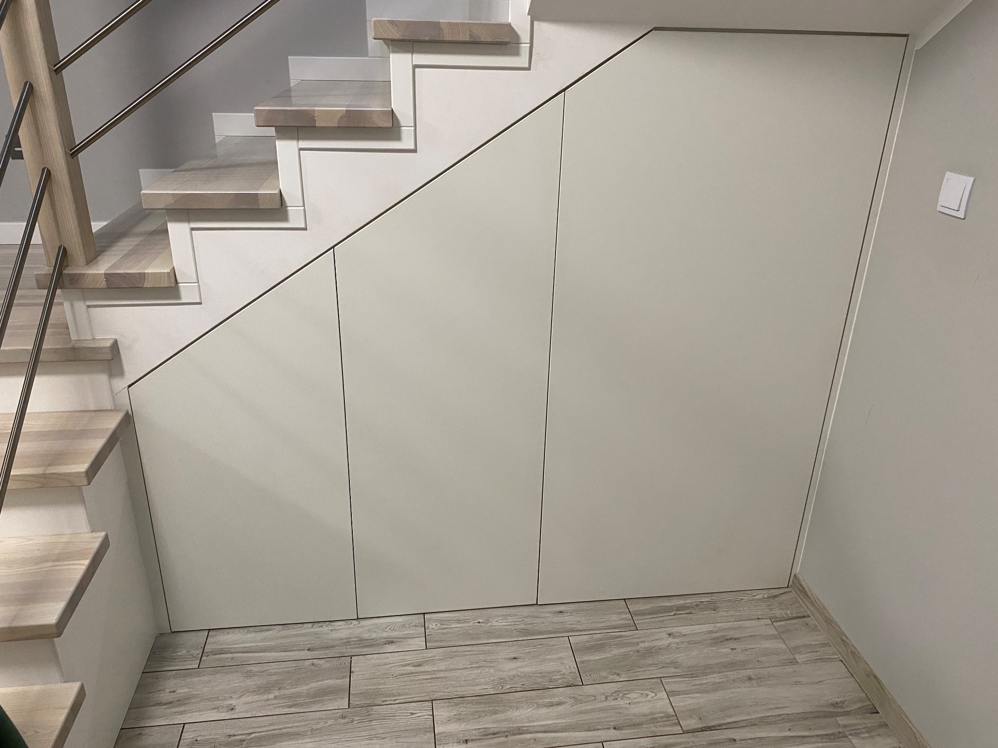 Biała szafa wnękowa pod schodami z drewnianymi stopniami, widoczna z boku, z fragmentem metalowej barierki i podłogą z płytek imitujących drewno.