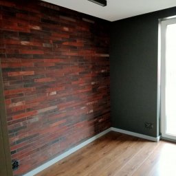 REM-TECH GRZEGORZ CICHY - Pokój po remoncie: ściana z cegły dekoracyjnej w odcieniach czerwieni i brązu, kontrastująca z gładką, ciemnoszarą ścianą, drewniana podłoga i nowoczesna lampa sufitowa.