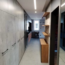 REM-TECH GRZEGORZ CICHY - Długi korytarz z szarą zabudową meblową po lewej stronie z czarnymi uchwytami, po prawej stronie półki z jasnego drewna na ścianie z cegieł, w tle widoczna kuchnia i pralka pod oknem.