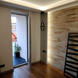 REM-TECH GRZEGORZ CICHY - Jasne wnętrze z drewnianą podłogą, ceglaną ścianą z oświetleniem punktowym, wiszącą lampą w kształcie infinity i otwartym balkonem z widokiem na zabudowę miejską.