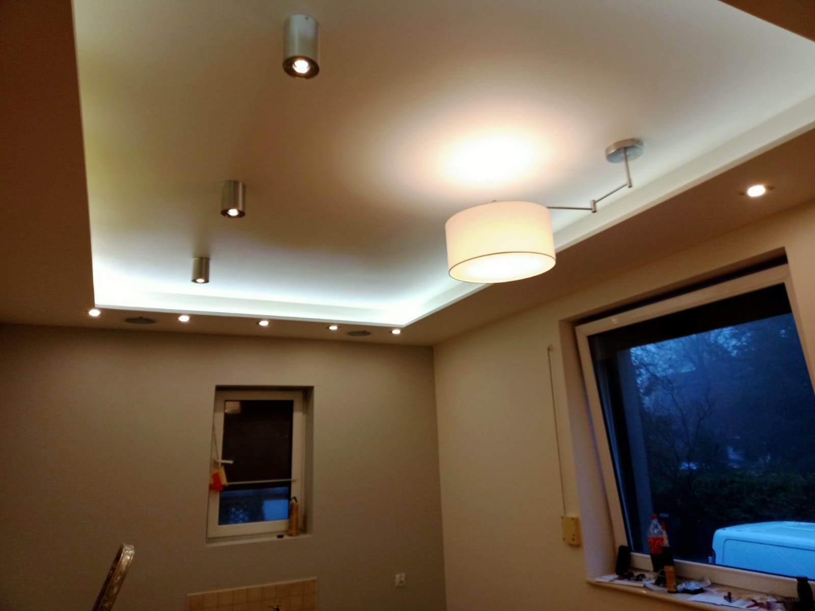 Wykończony sufit podwieszany z oświetleniem punktowym LED i lampą wiszącą na wysięgniku, widok z dołu na świeżo pomalowane ściany i okno z widokiem na zewnątrz.