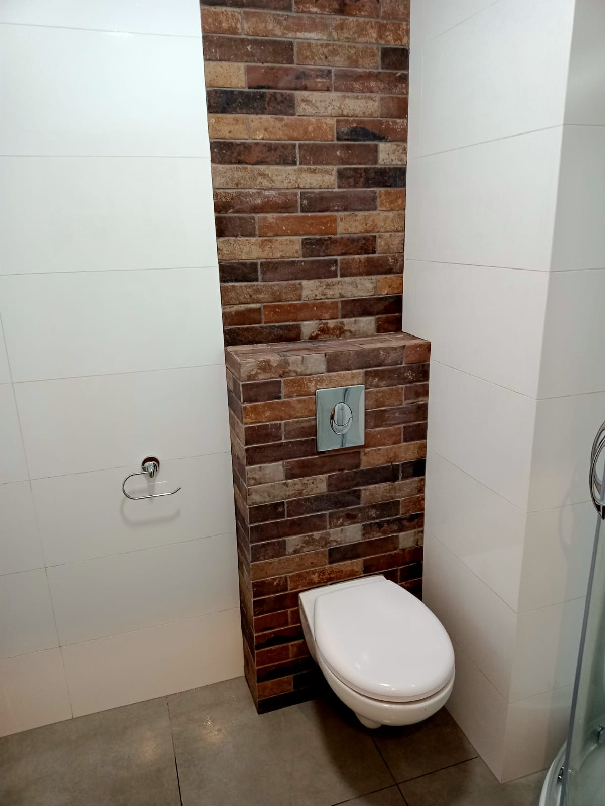 Nowoczesna toaleta podwieszana z obudową z cegły klinkierowej, białe płytki na ścianach i szara podłoga.