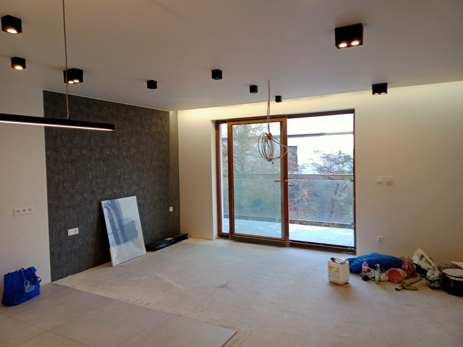Wnętrze w trakcie prac wykończeniowych: surowa podłoga, ściana z tapetą strukturalną, nowoczesne oświetlenie sufitowe i narzędzia malarskie przy oknie balkonowym.