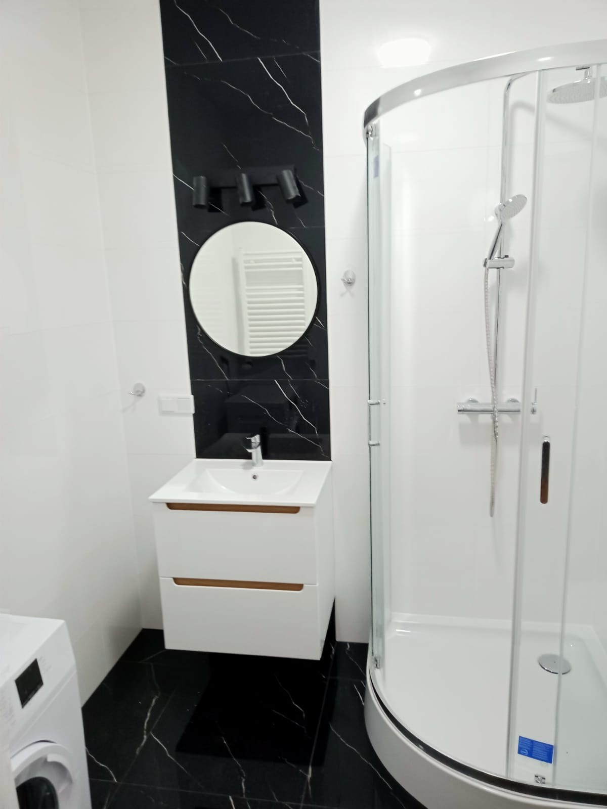 Nowoczesna łazienka z czarno-białymi płytkami imitującymi marmur, okrągłym lustrem z oświetleniem punktowym, białą szafką pod umywalkę z drewnianymi akcentami i kabiną prysznicową z zaokrąglonymi...