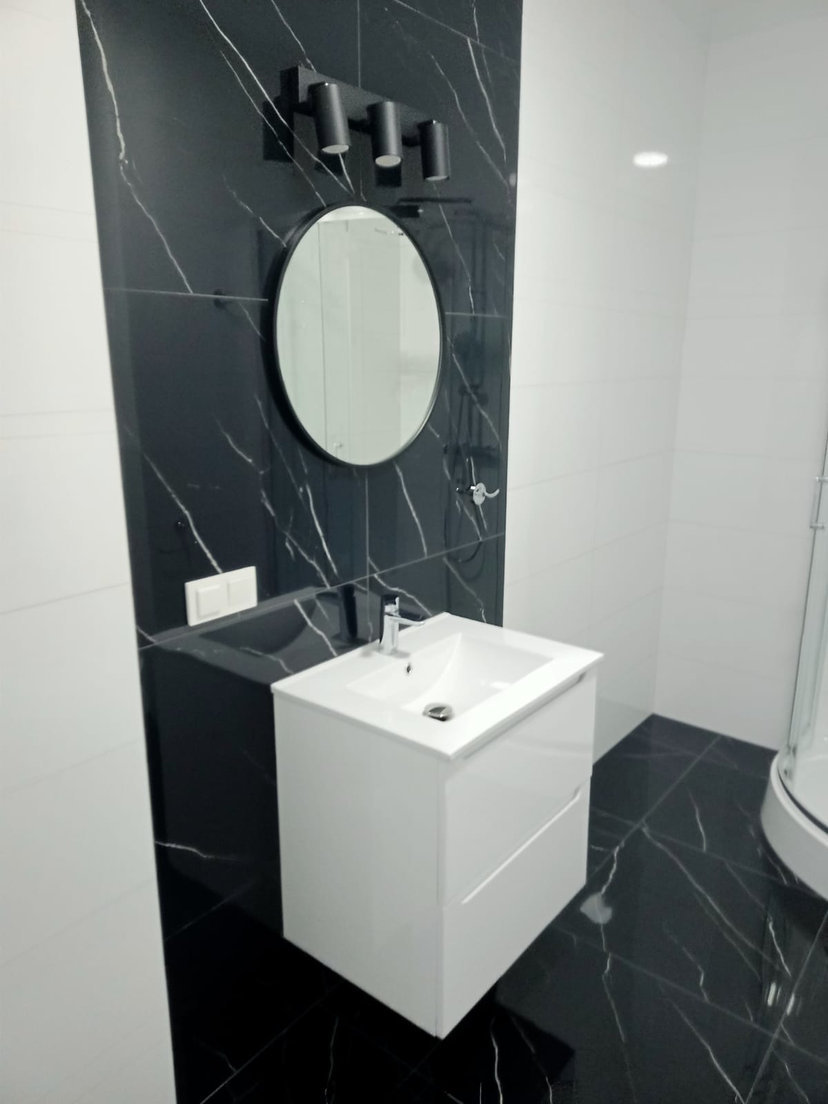 Nowoczesna łazienka z czarno-białymi płytkami imitującymi marmur, okrągłym lustrem pod trzema punktowymi lampami i białą szafką umywalkową z chromowaną baterią.