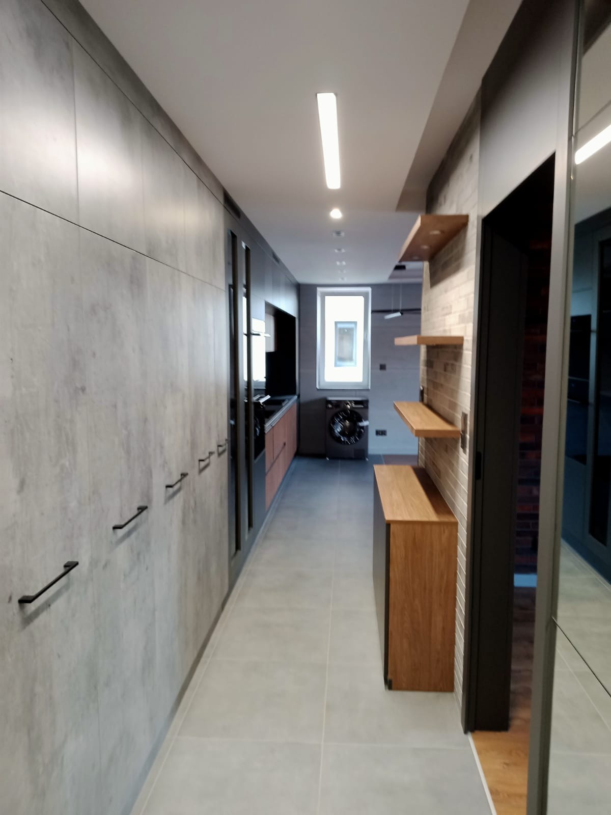 Długi korytarz z szarą zabudową meblową po lewej stronie z czarnymi uchwytami, po prawej stronie półki z jasnego drewna na ścianie z cegieł, w tle widoczna kuchnia i pralka pod oknem.