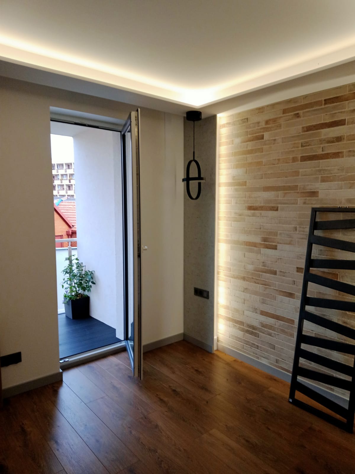 Jasne wnętrze z drewnianą podłogą, ceglaną ścianą z oświetleniem punktowym, wiszącą lampą w kształcie infinity i otwartym balkonem z widokiem na zabudowę miejską.