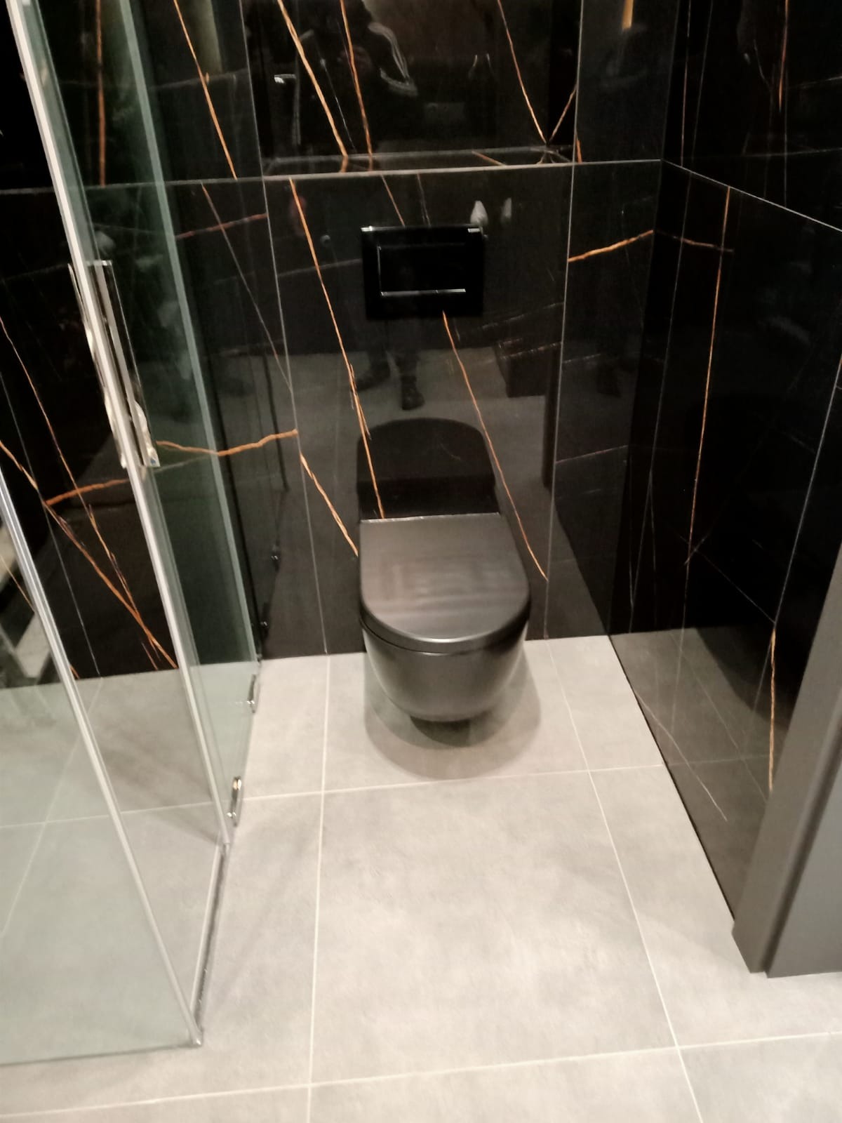 Nowoczesna toaleta w czarnym kolorze, wisząca na tle czarnych płytek z żyłkowaniem w kolorze złota, szare płytki podłogowe, kabina prysznicowa ze szklanymi drzwiami.